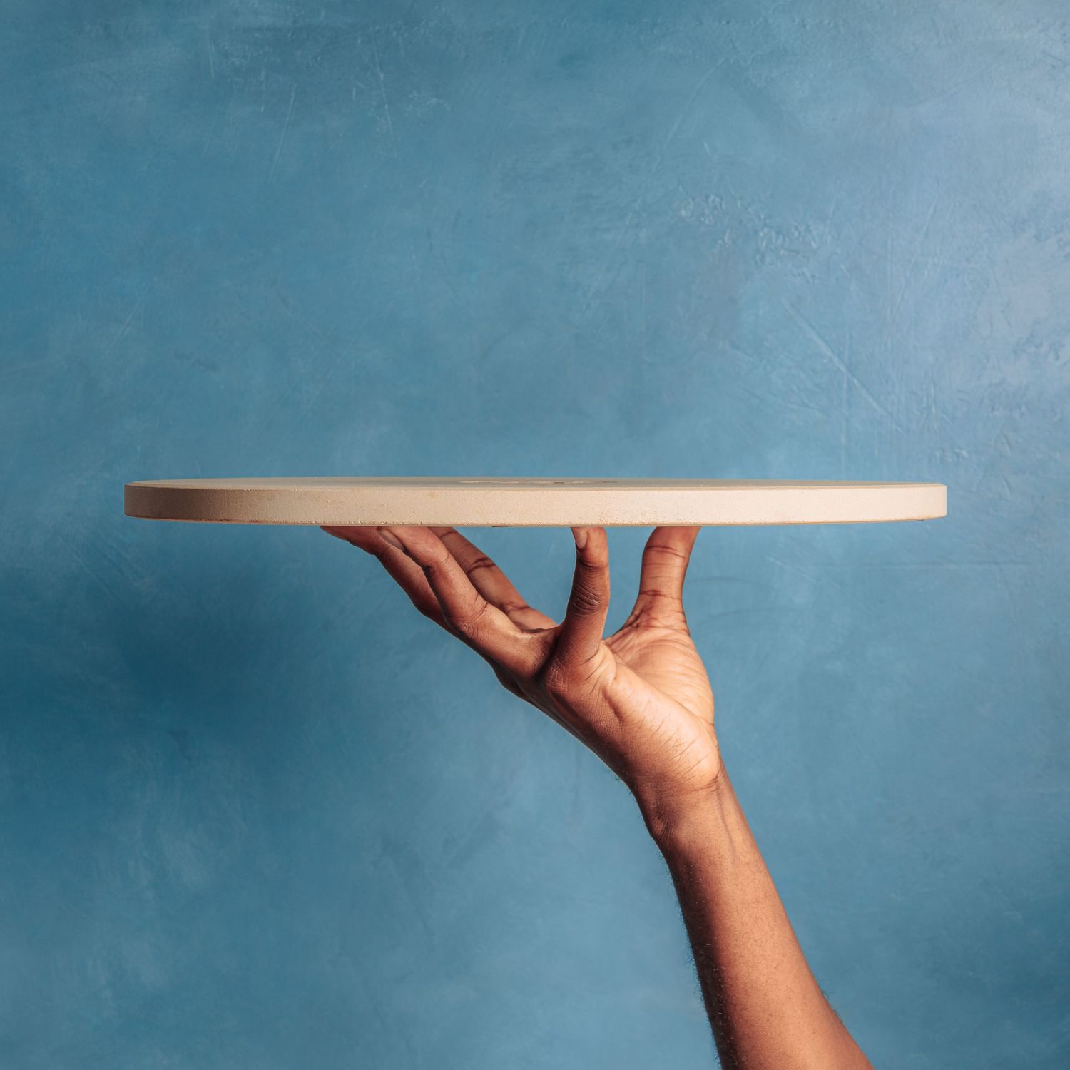 Hand holding up a round white serving platter against a blue background.