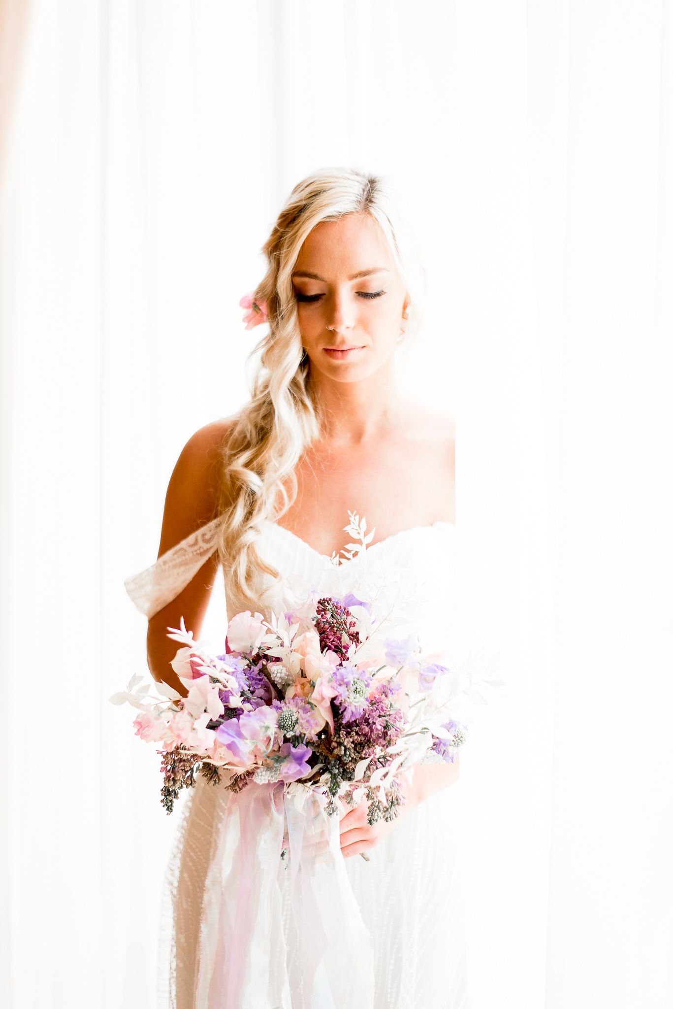 blonde bride with braided hair next to a window looking at purple and white wedding bouquet at The Center Cincinnati