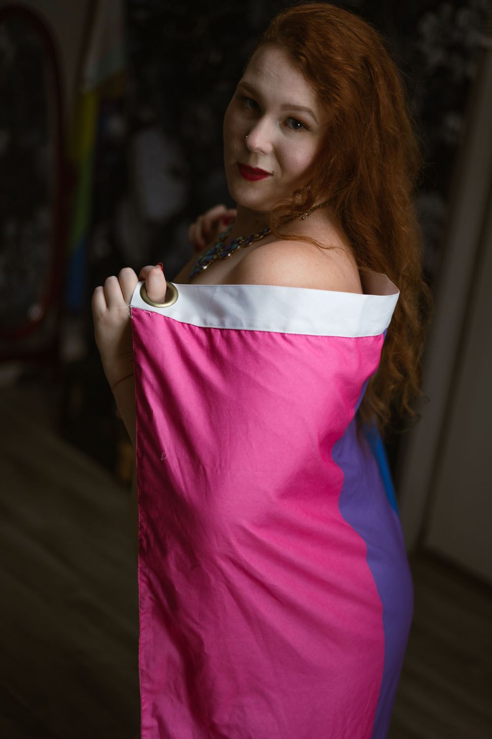 curvy woman wrapped in only a bisexual pride flag looks over her shoulder at the viewer.