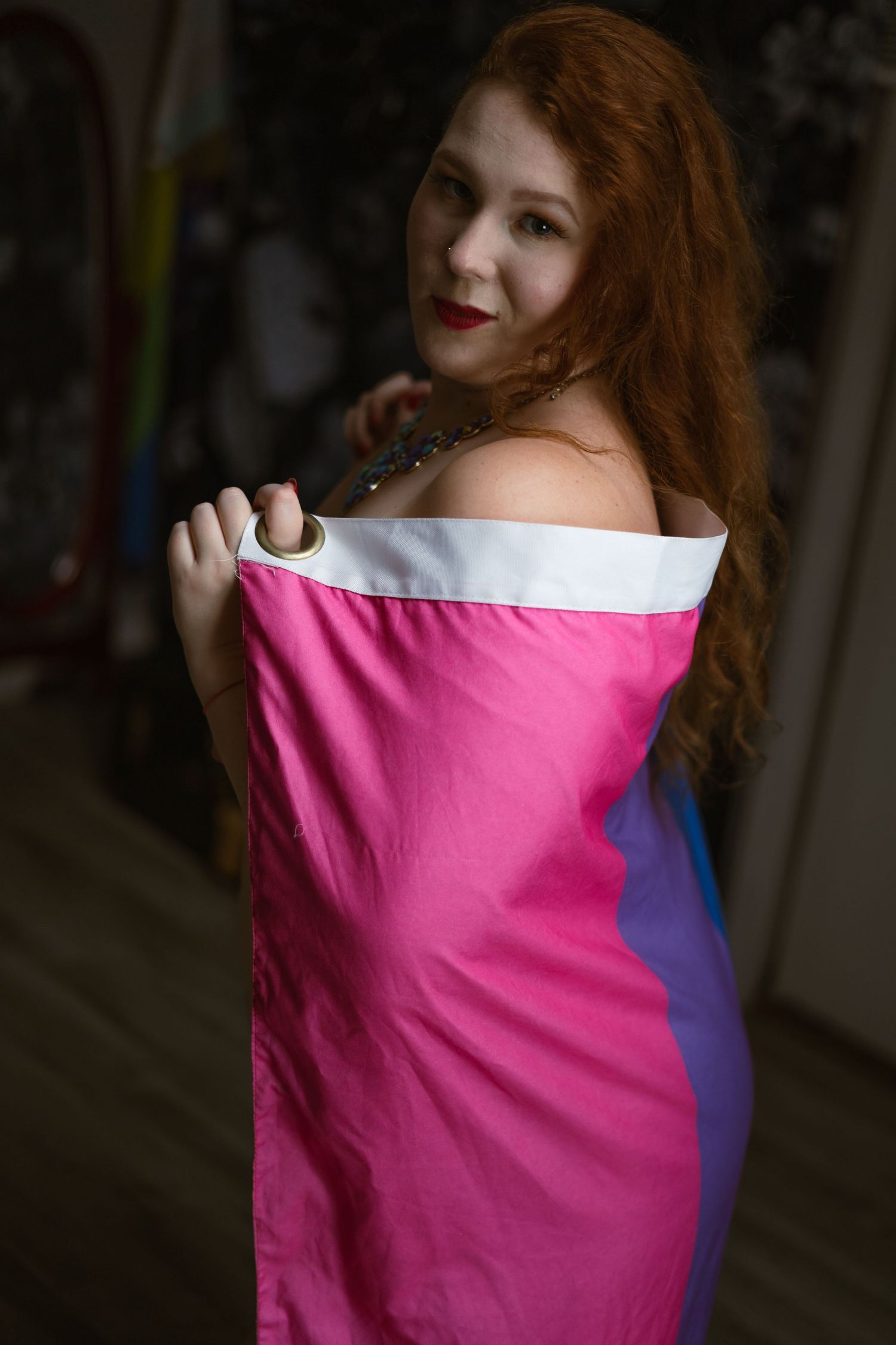 curvy woman wrapped in only a bisexual pride flag looks over her shoulder at the viewer.