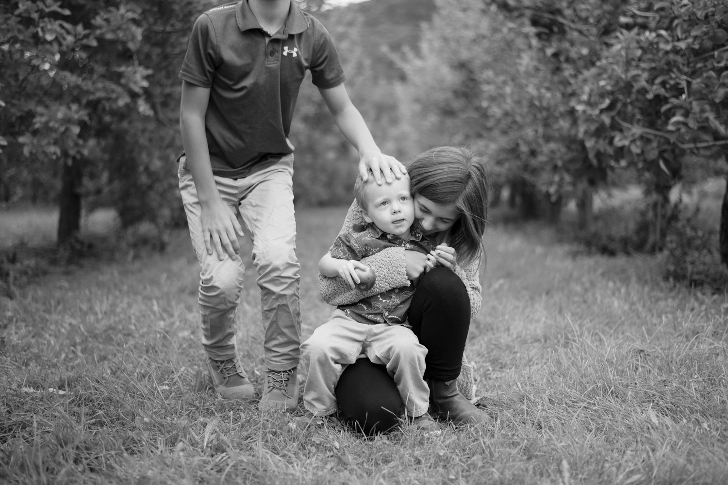 an older sibling hugs her younger brother as the older brother walks away and gives him a pat on the head