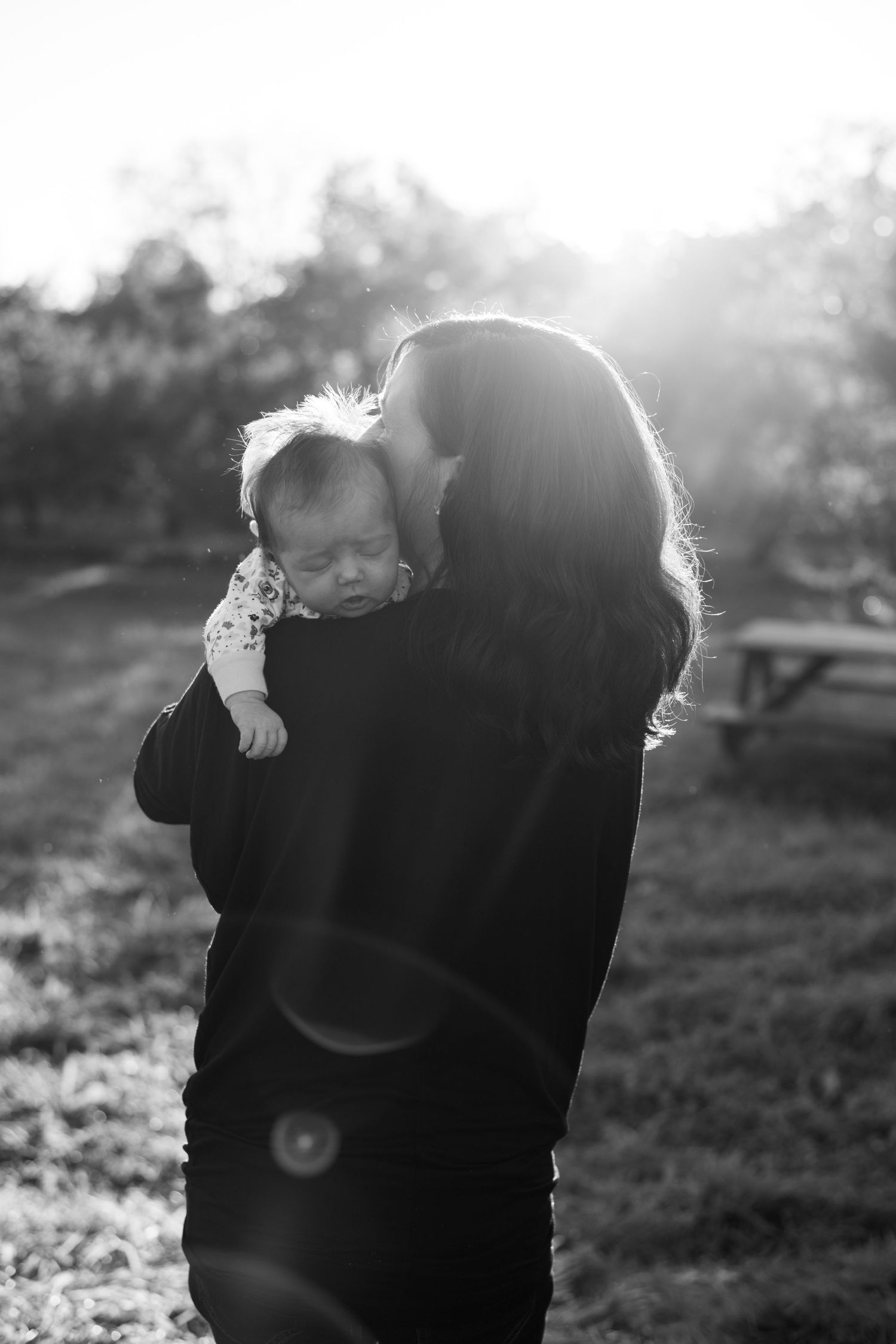 a mother embraces her newborn baby over her shoulder as the sun sets behind them in this black and white image