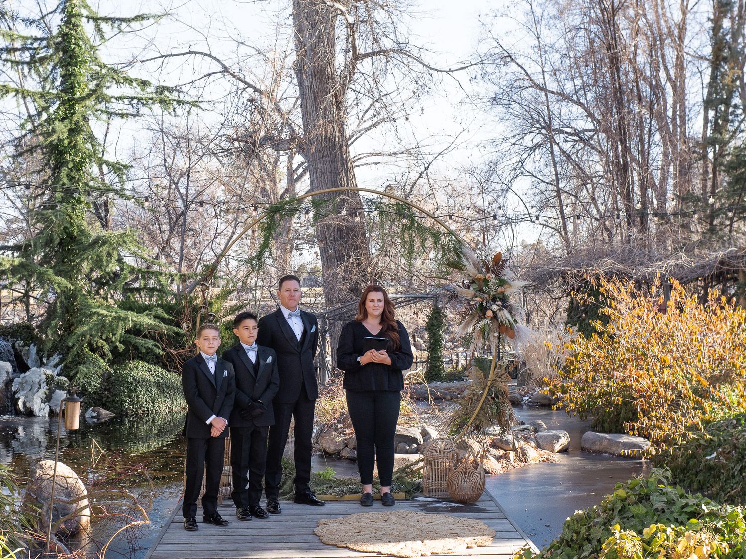A small group stands together on a garden path surrounded by evergreen trees and autumn foliage on a cloudy day, waitng for the bride.