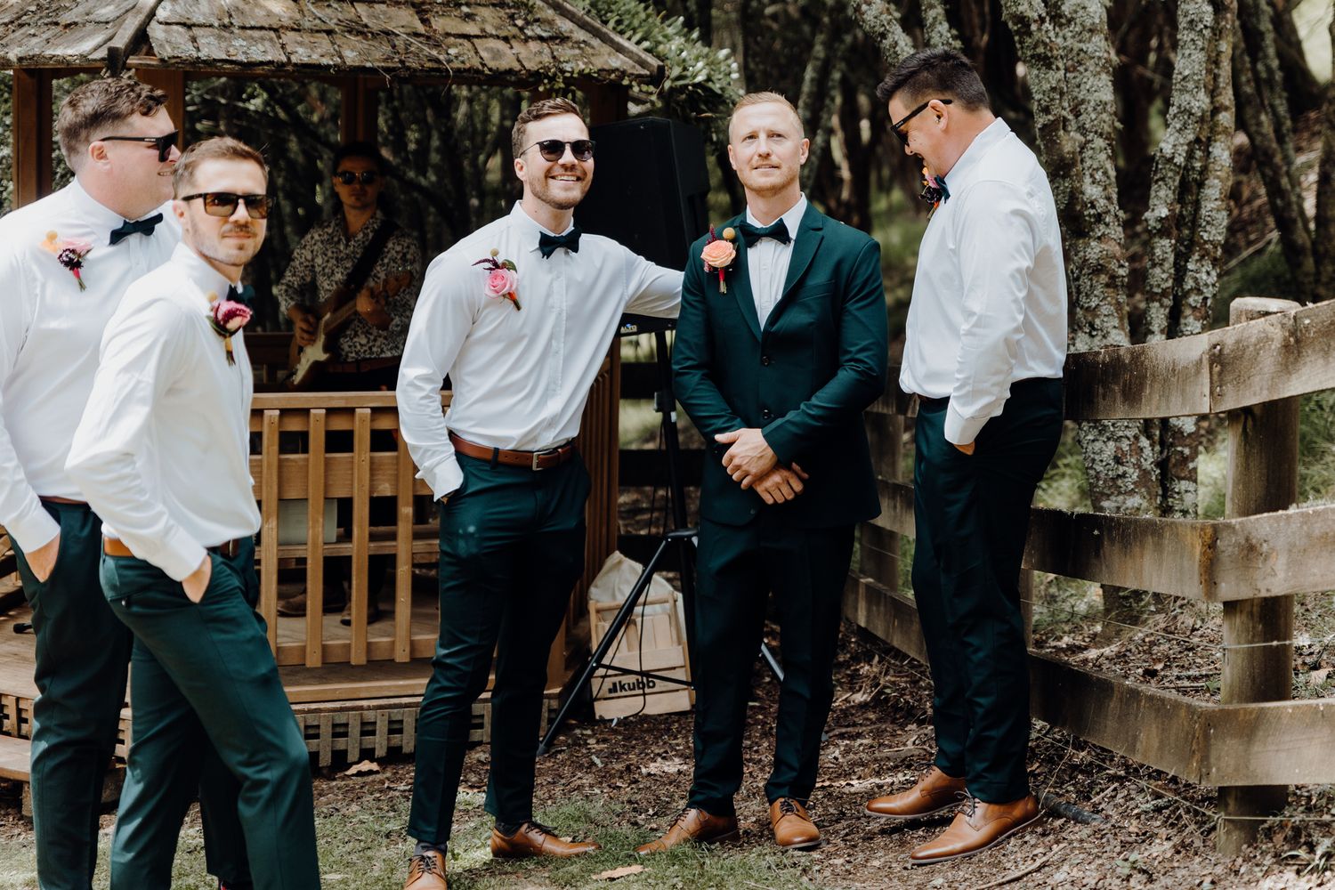 Wedding party in formal attire gathered near a rustic wooden gazebo.