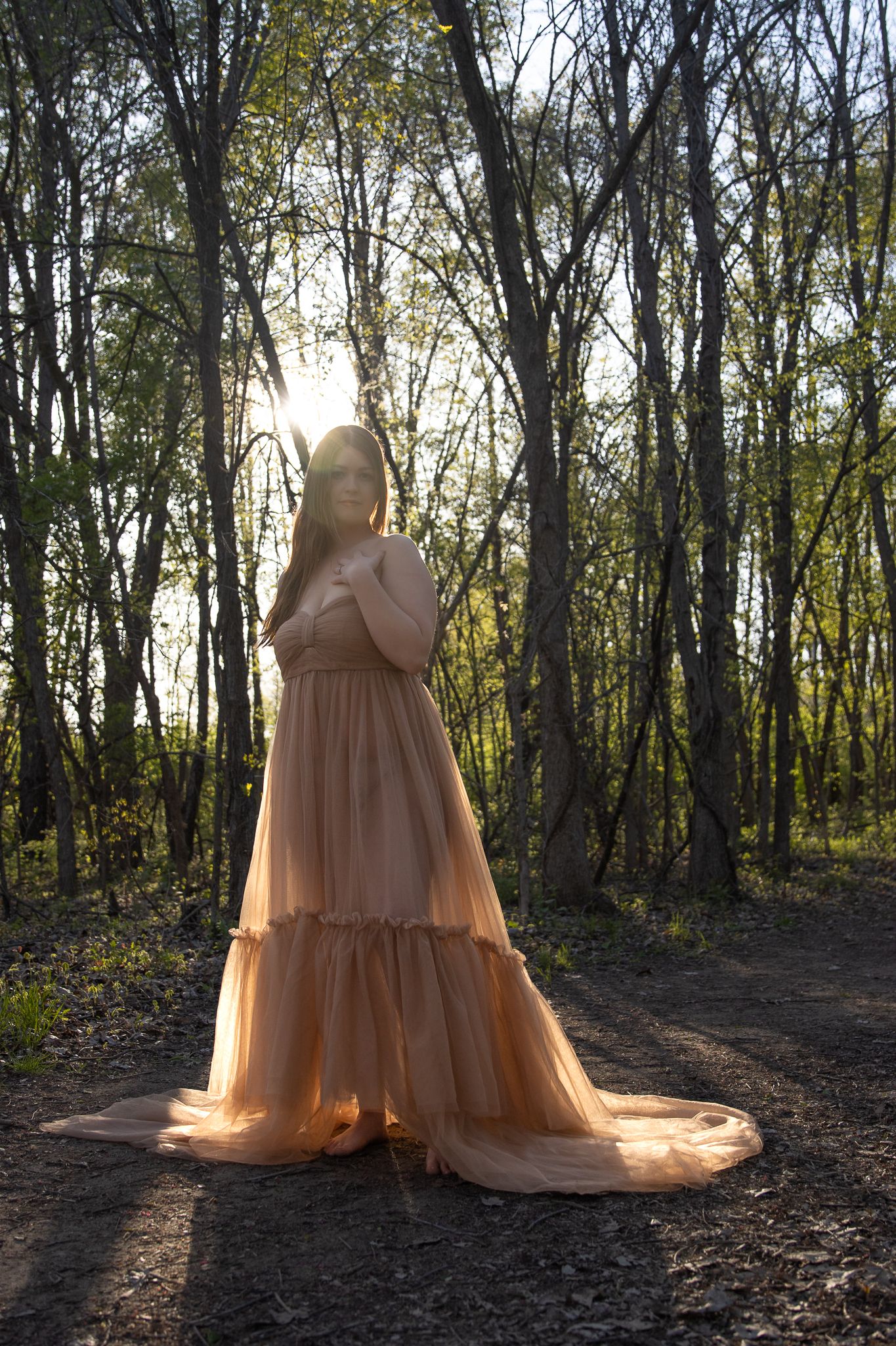 A dreamy peach-colored maternity dress silhouetted against sunset in a wooded setting.
