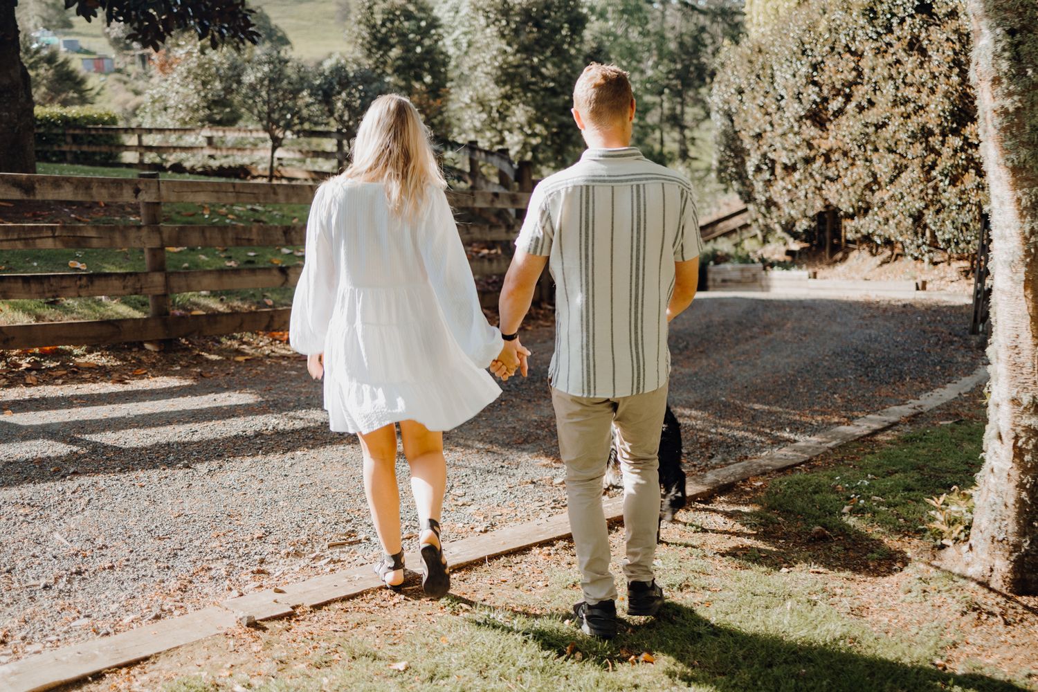 A couple walks hand in hand down a tree-lined gravel path on a sunny day.
