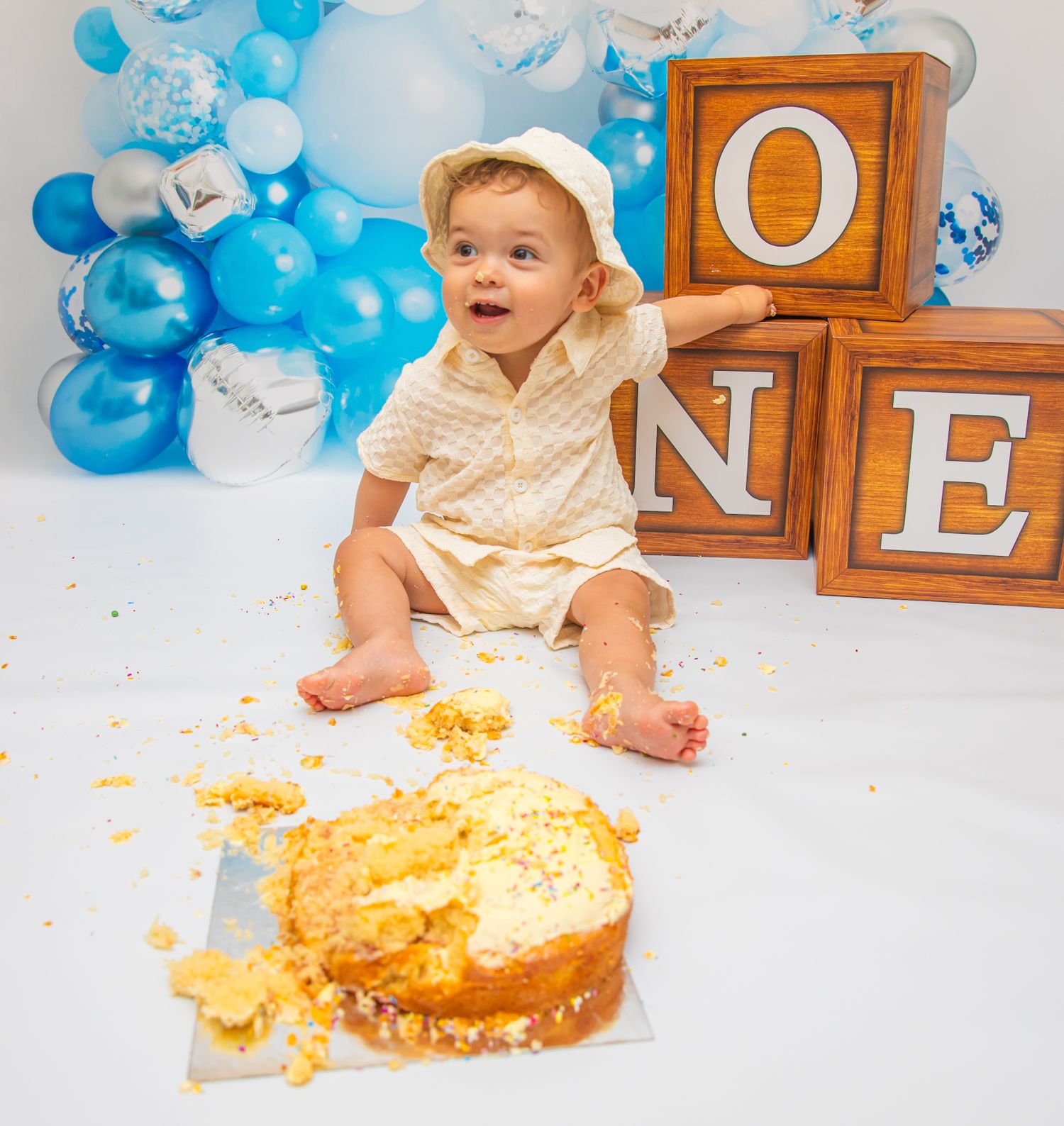 Cake smash photography session with wooden letter blocks spelling ONE against blue balloon display.