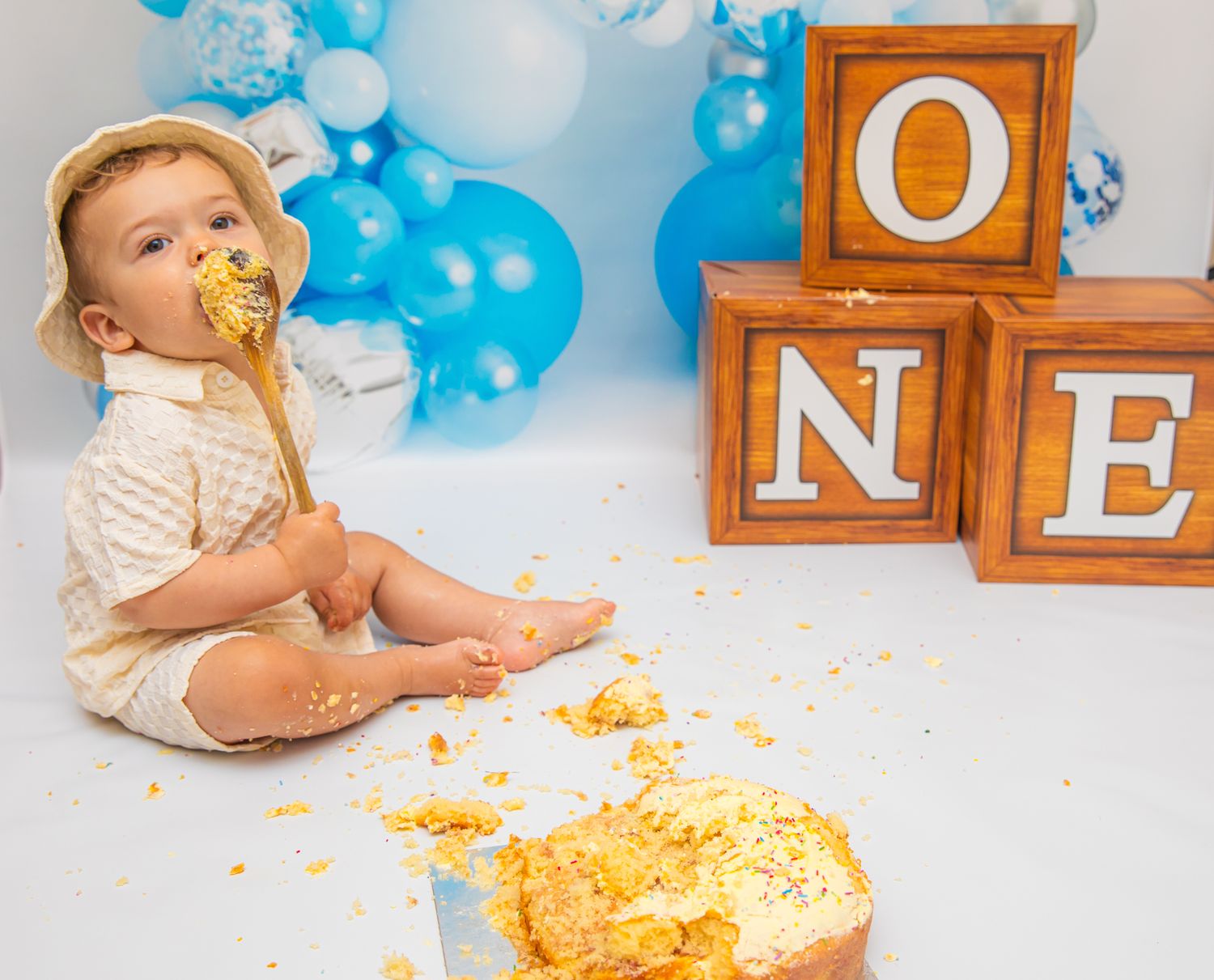 Baby enjoying cake smash session with wooden ONE blocks and blue balloon backdrop.