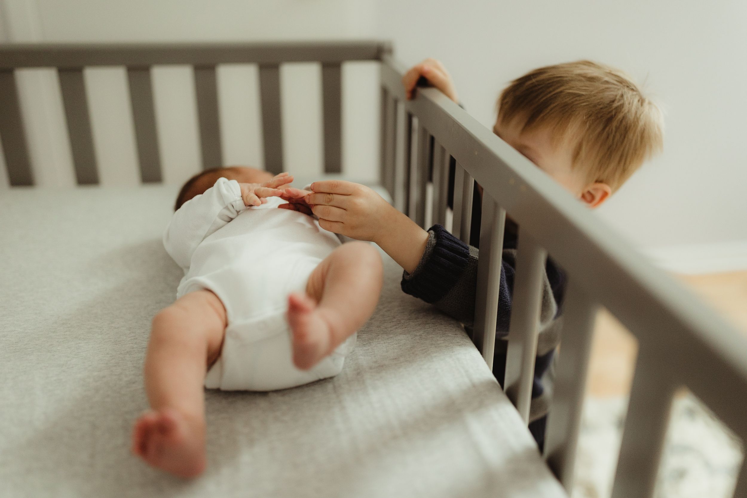 Newborn in a crib while toddler brother reaches in and holds her hand. Photo by newborn photographer, Elizabeth Pachniak