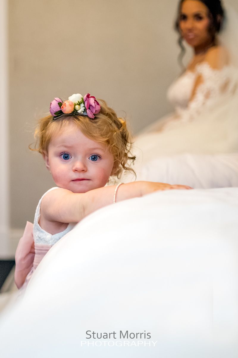 little flower girl looks straight at the camera while leaning on the bed bride sits on the bed in the background