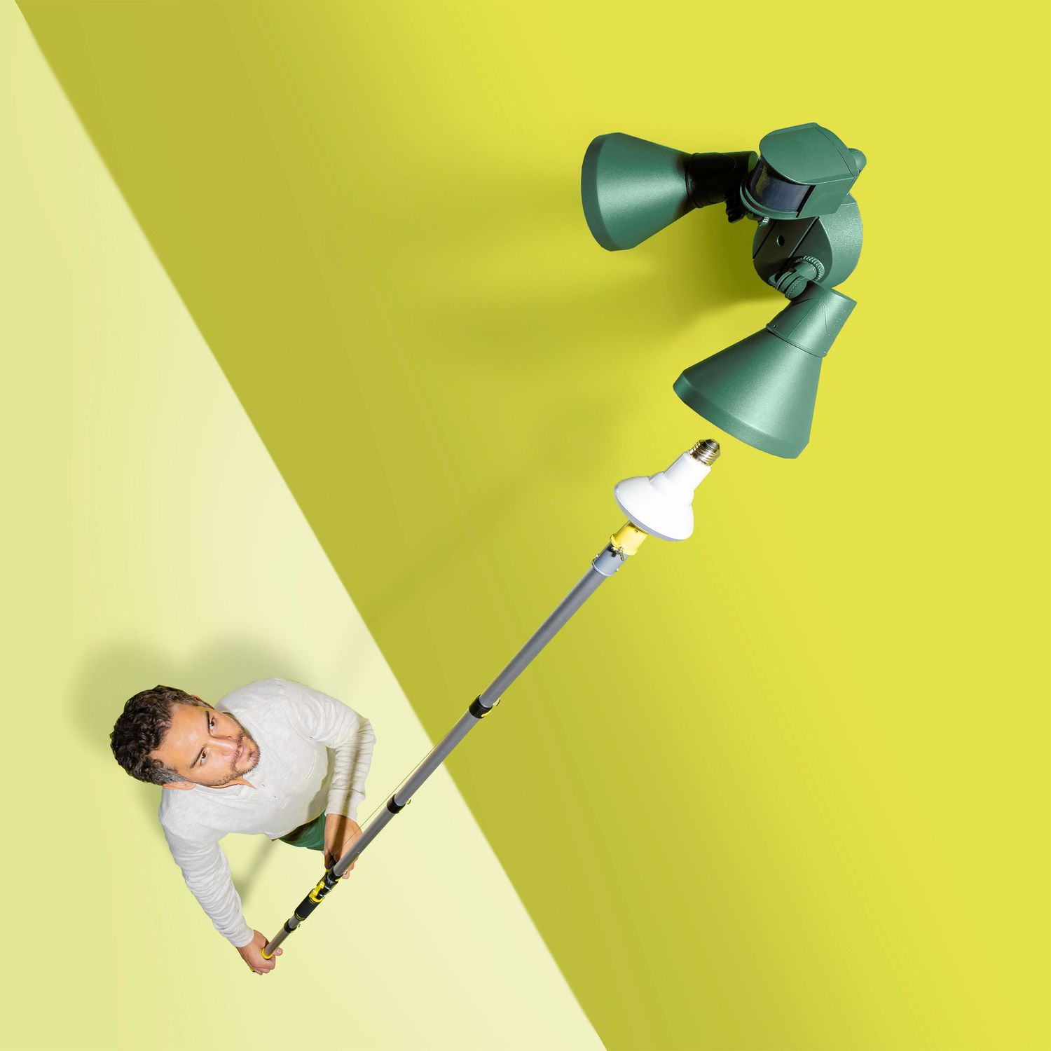 Minimalist photo of extending pole reaching wall-mounted light fixture against yellow backdrop.