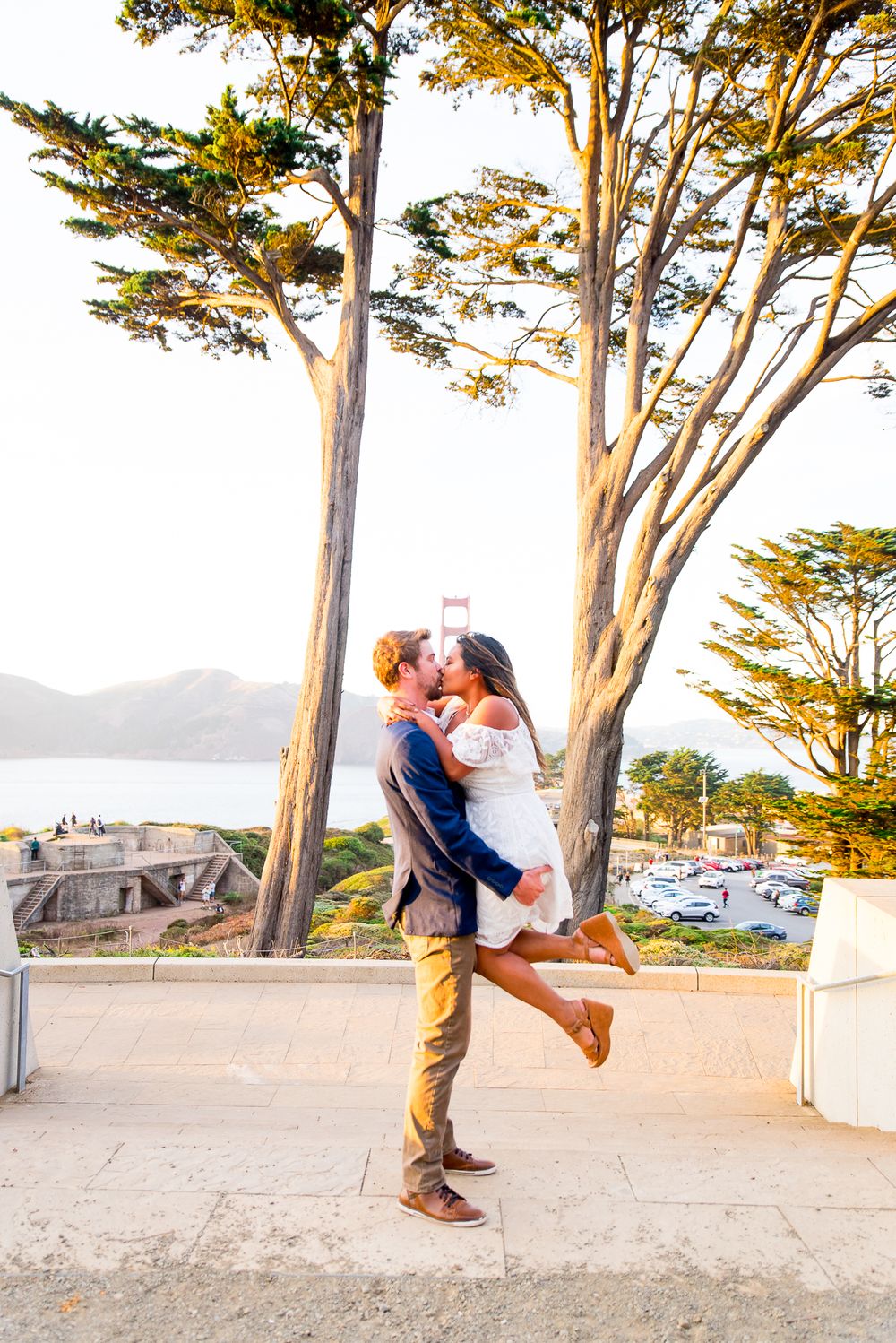 man picking up woman in white dress kissing in front of Golden Gate Bridge engagement pictures