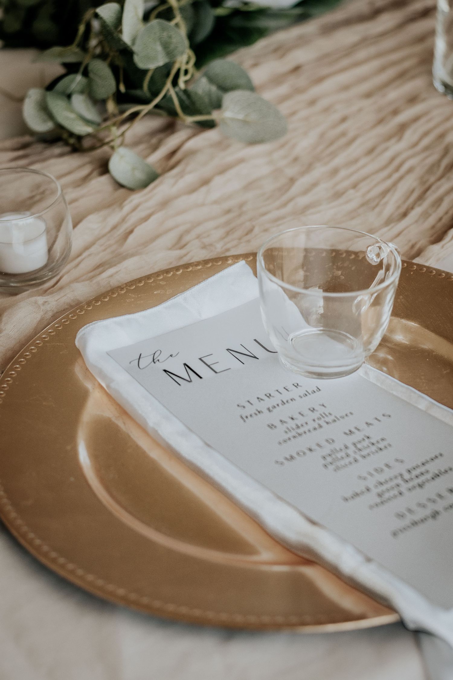 Elegant wedding place setting with gold charger plate, menu card and eucalyptus decor on textured tablecloth.