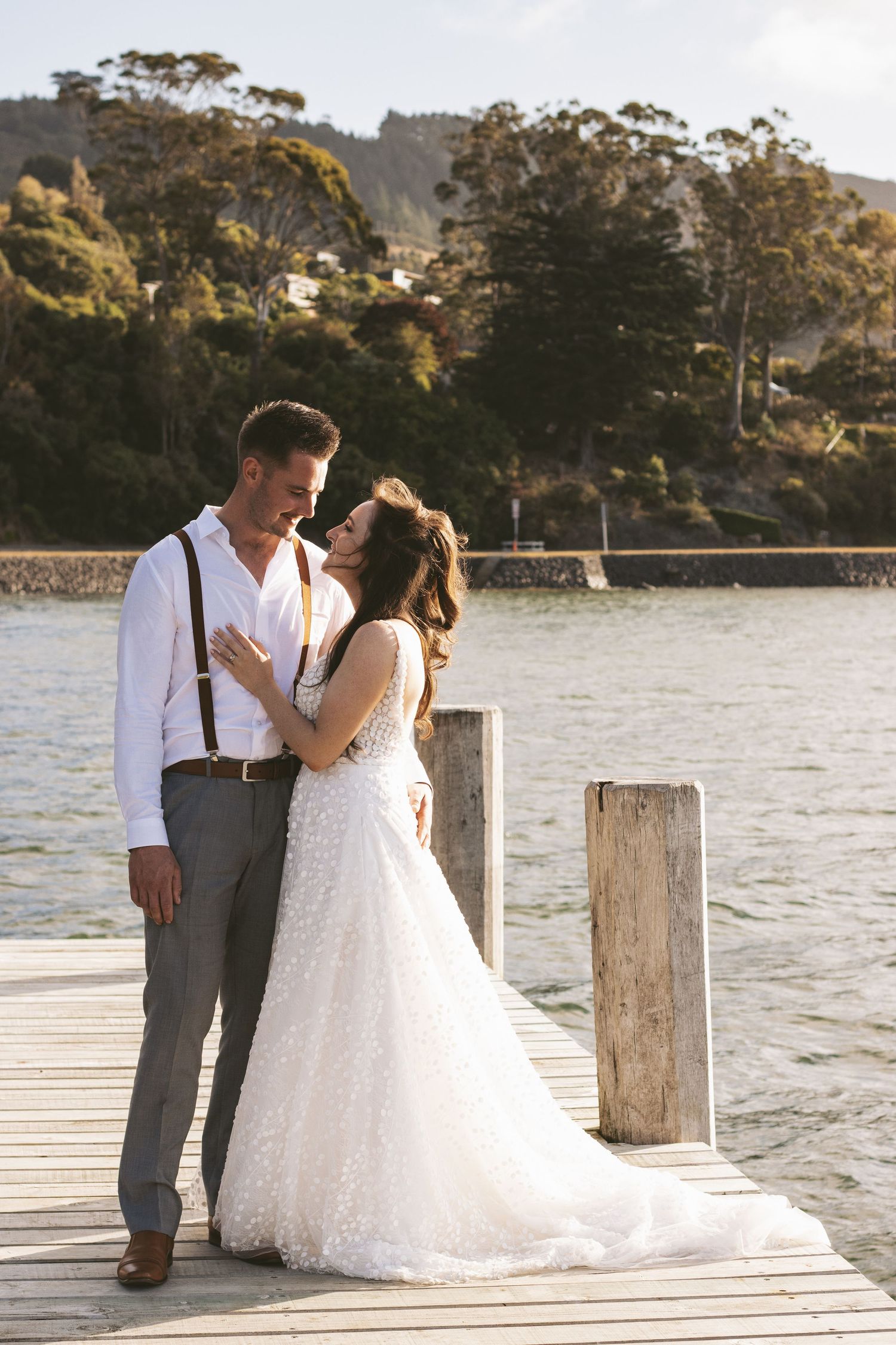 Wedding couple embracing on wooden dock by the water with scenic mountain backdrop.