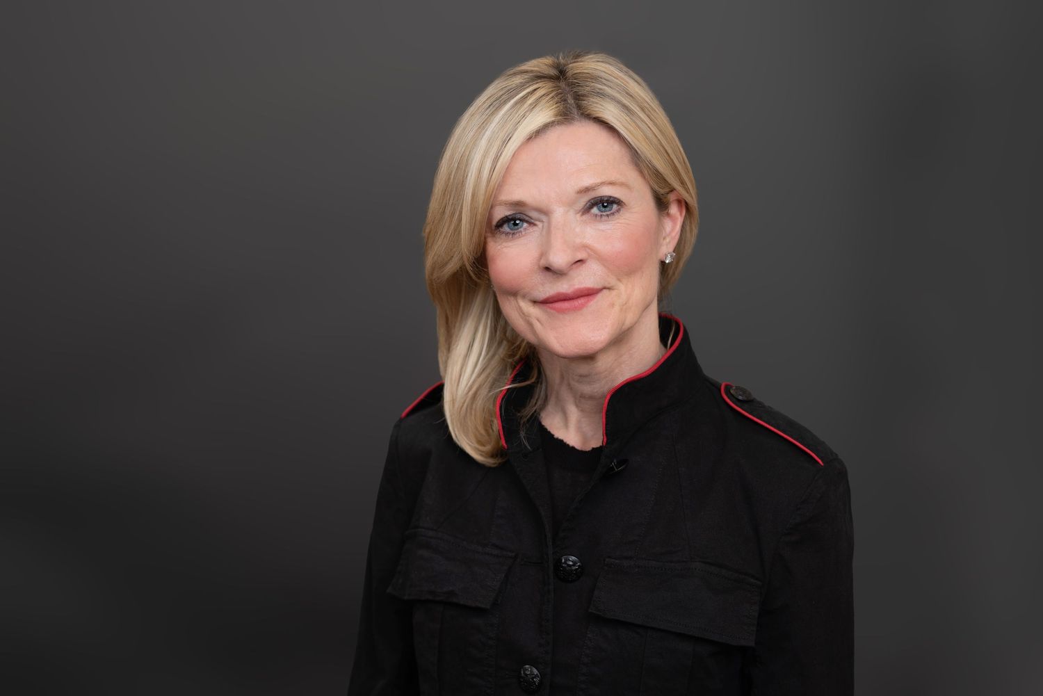 Professional headshot of woman in black jacket against dark background.