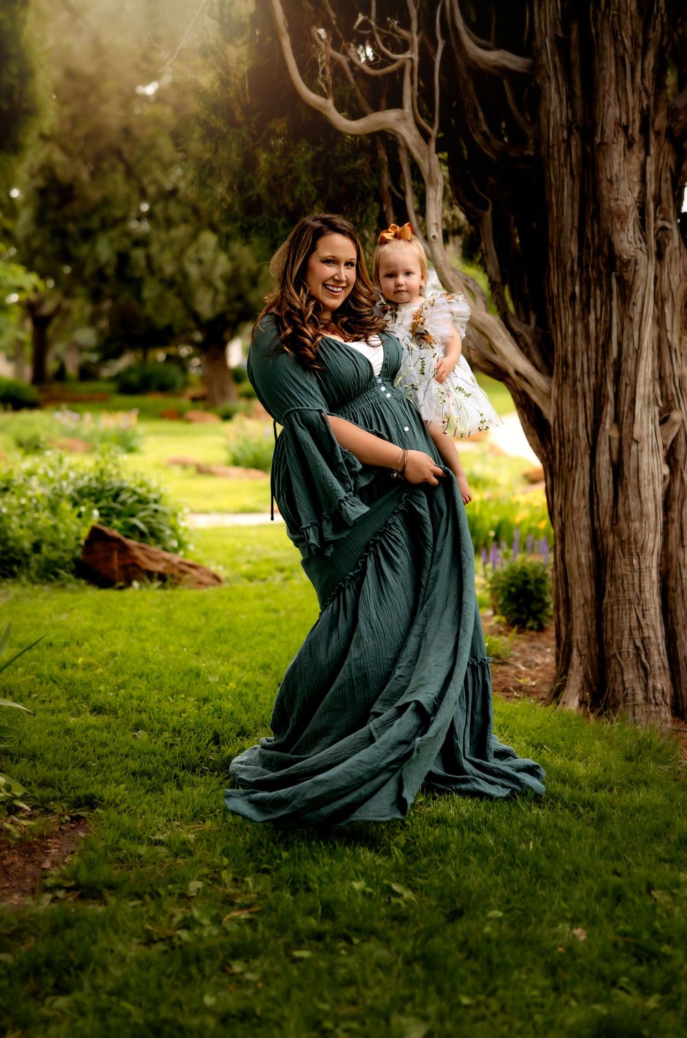 Family portrait in a park with flowing emerald green dress against a large tree trunk in warm natural lighting.