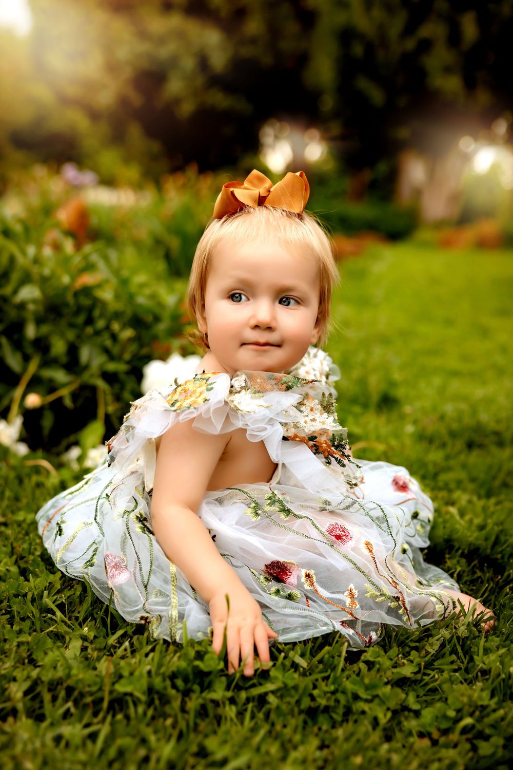 A small child in a floral dress and orange bow sits on green grass during golden hour lighting.