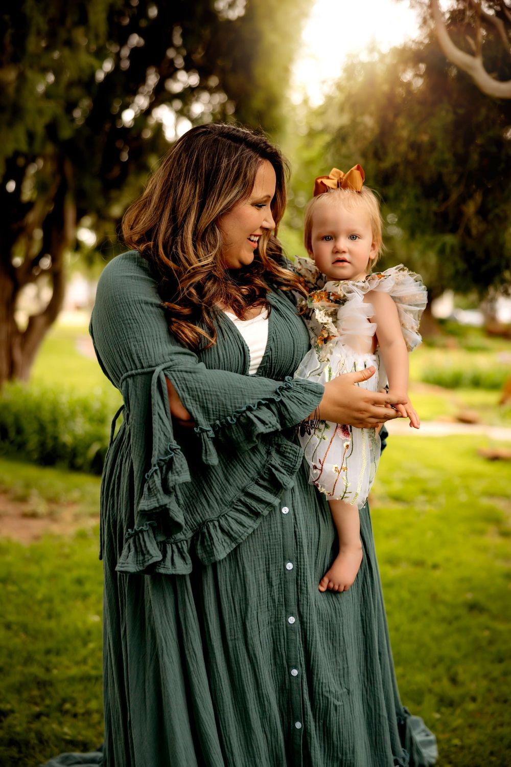 A tender moment captured outdoors at sunset with a woman in a flowing green dress holding a baby in a ruffled outfit.