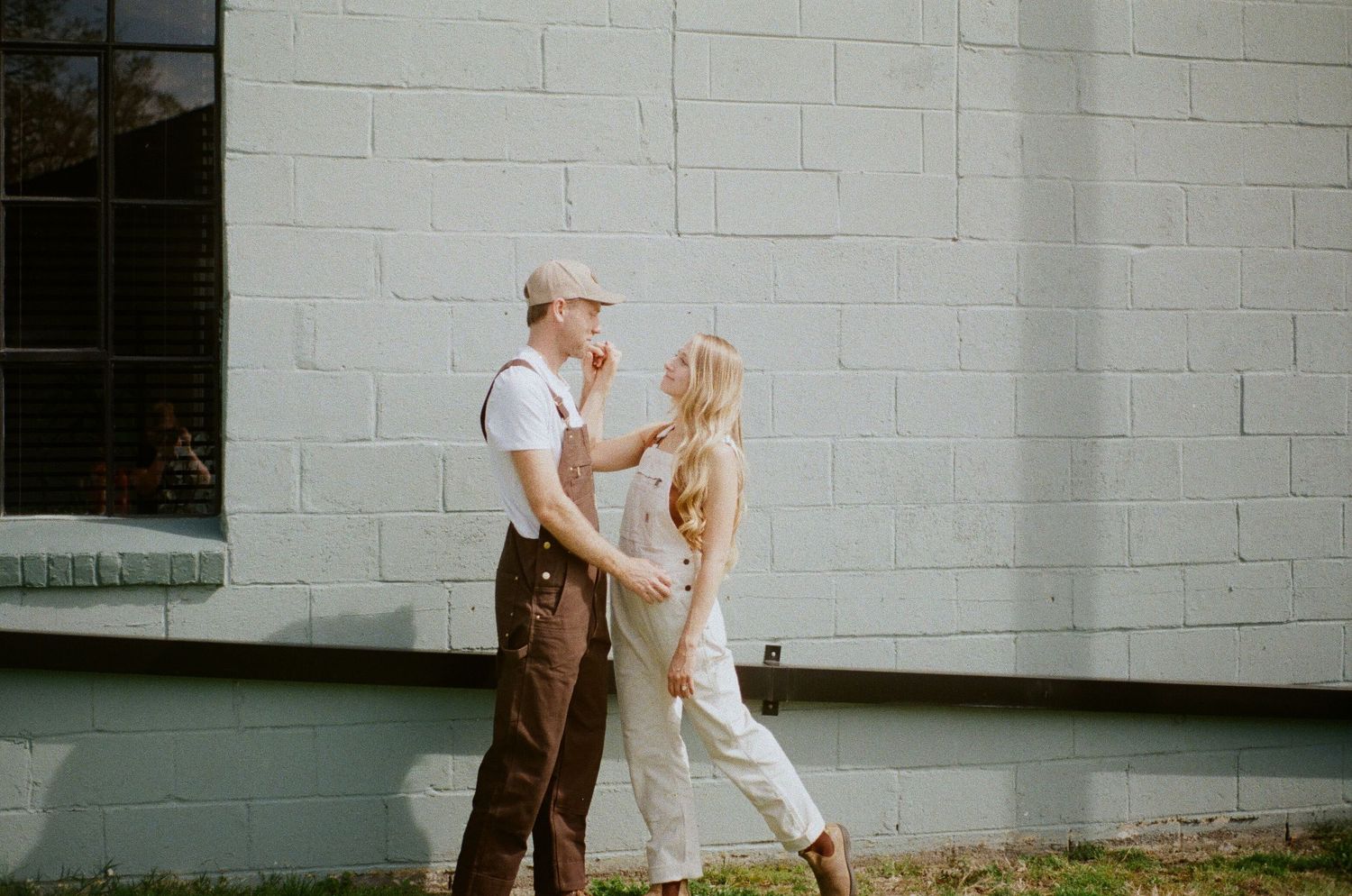 A couple dances playfully together against a white brick wall on a sunny day.