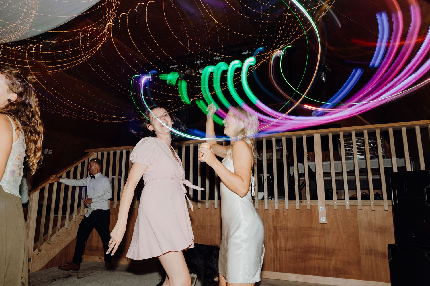 Dancers celebrate under swirling multicolored neon lights at an indoor wedding reception.