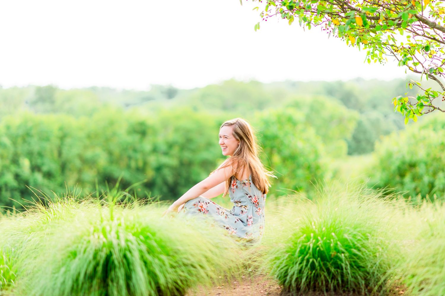 young woman sitting in field of tall light green grass under overcasts sky laughing for Chicago senior pictures