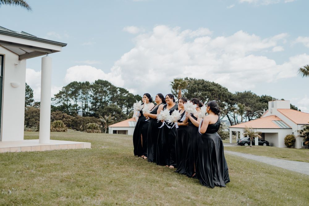 Wedding party in black dresses poses on grassy hill with white building and palm trees.