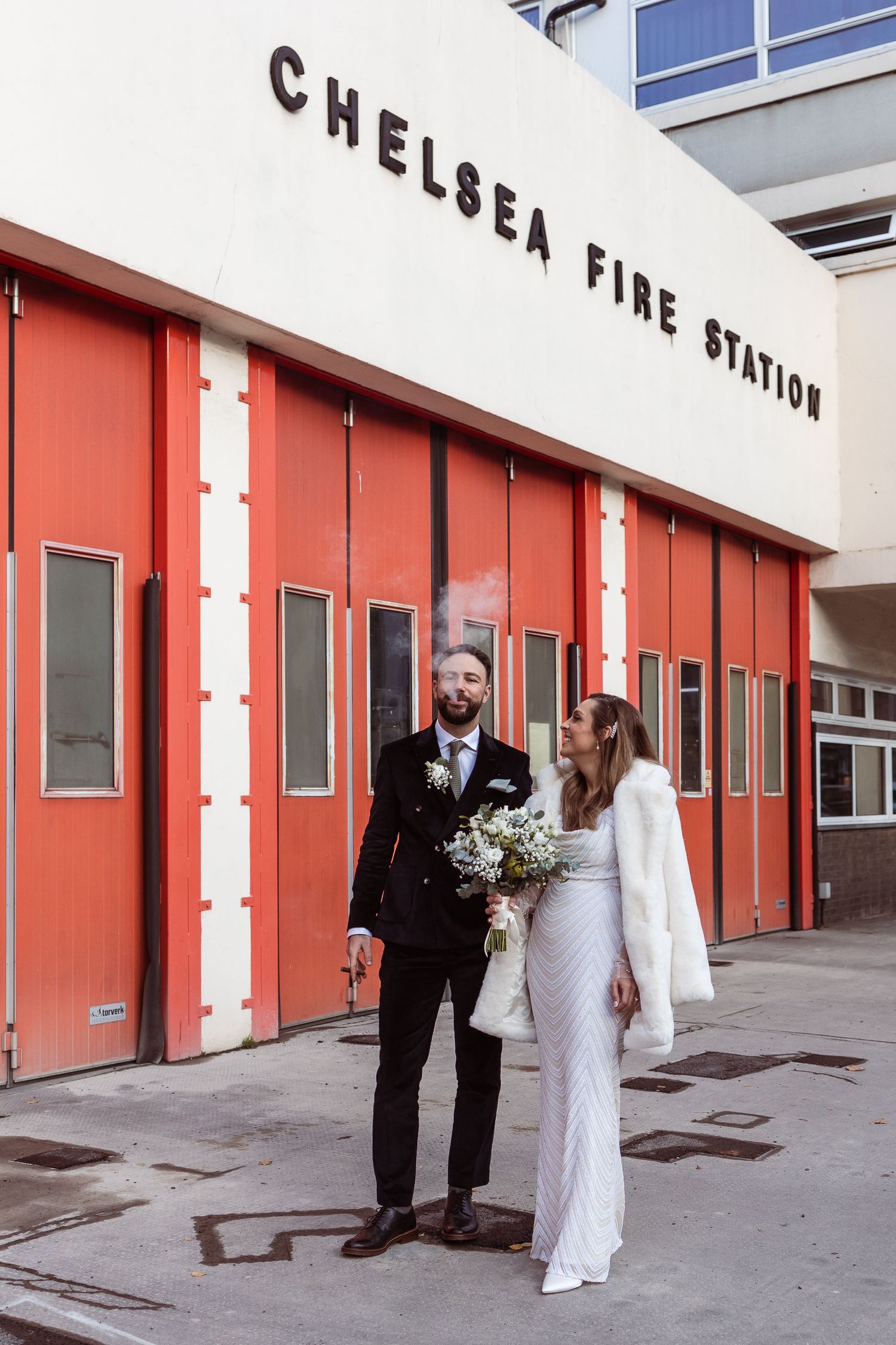 Couple portrait of a bride and groom, outside Chelsea Fire Station, smoking a cigar.