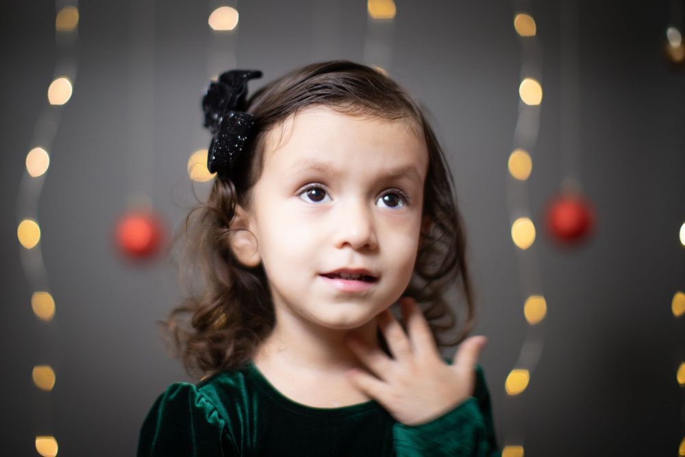 Child in green velvet dress poses against bokeh lights and holiday decorations.