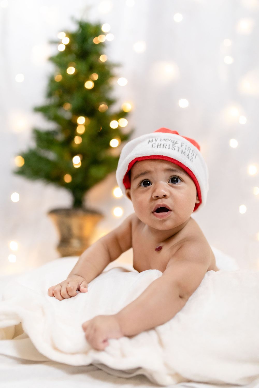 Baby in First Christmas hat reaches toward camera with decorated tree in background.