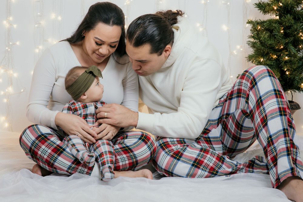 Family cuddles together on white blanket wearing matching plaid pajamas with Christmas lights in background.