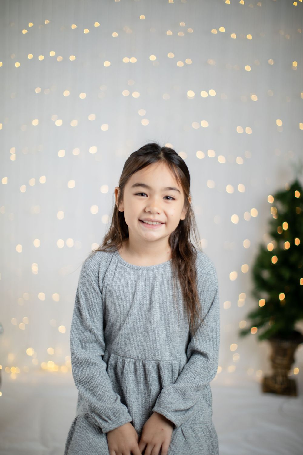 Young girl in grey dress poses against sparkly holiday backdrop with Christmas tree.