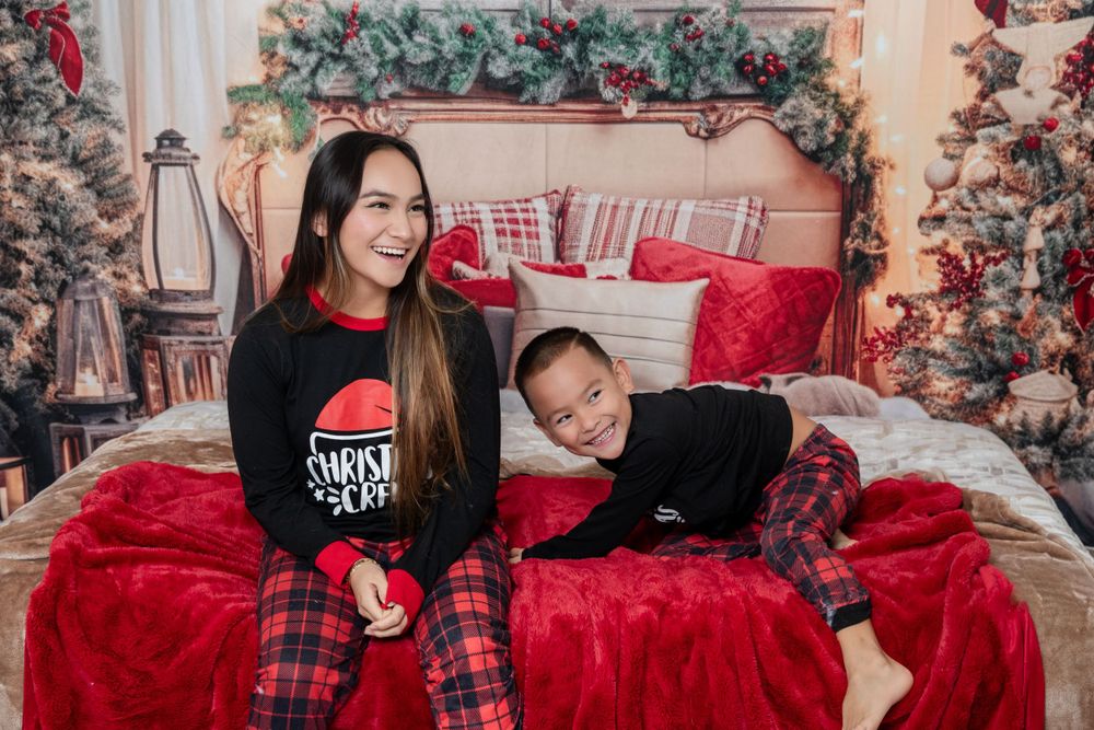 Mother and child wearing matching Christmas pajamas pose together on a red velvet bed with holiday decorations.