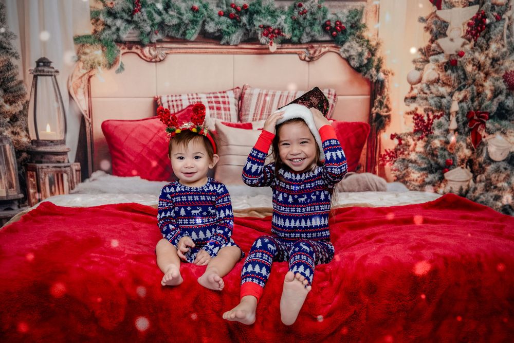 Two children in matching blue winter pajamas sit on a red bed with festive holiday decorations.