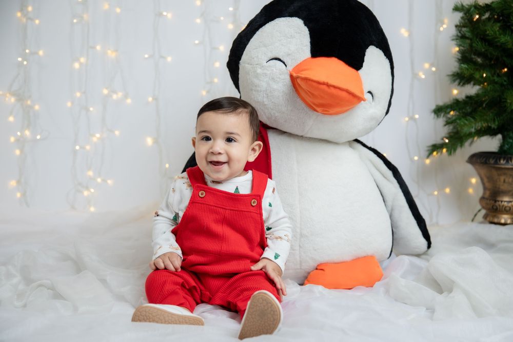 Baby in red outfit sits next to large plush penguin with Christmas tree in background.