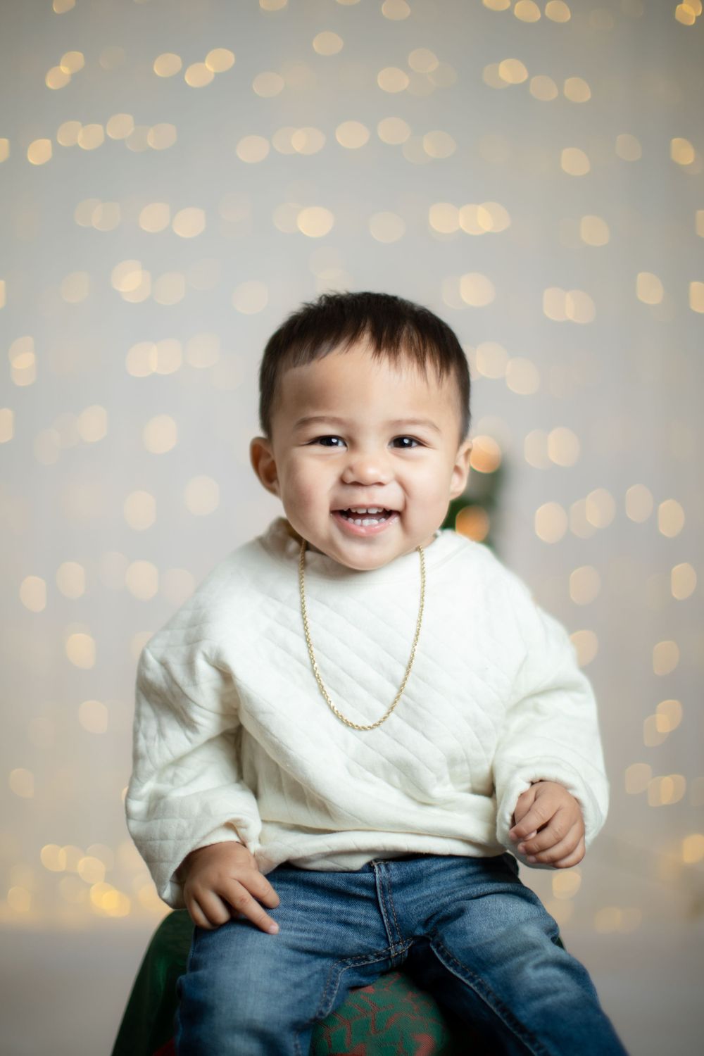 Baby in white sweater smiling with sparkly bokeh lights in background during holiday photoshoot.