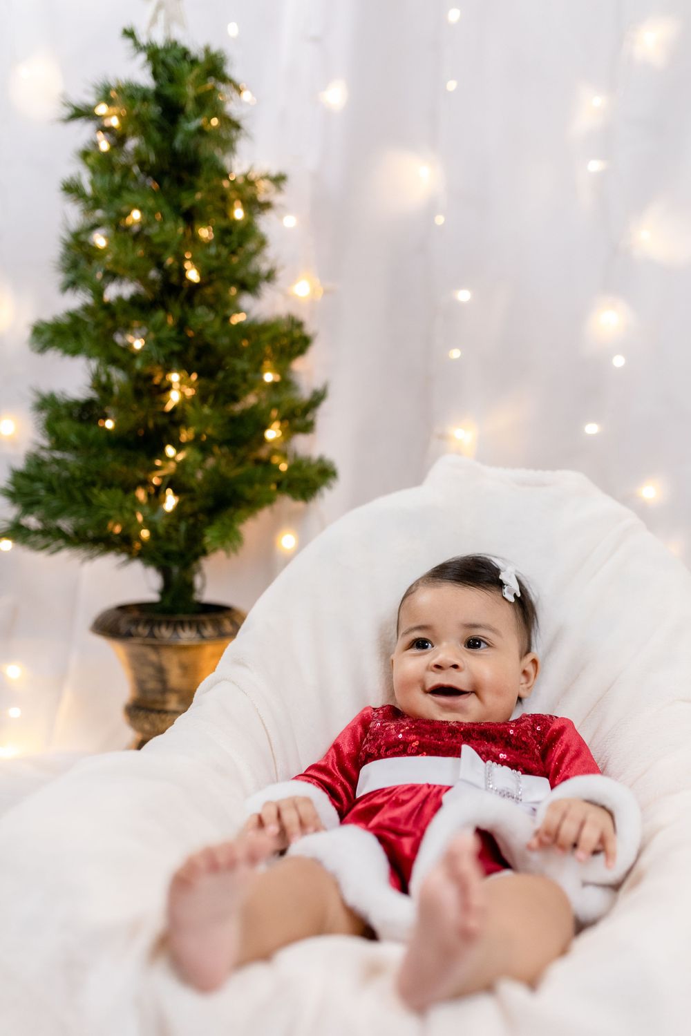 Baby in red festive outfit lying on white chair next to decorated Christmas tree.