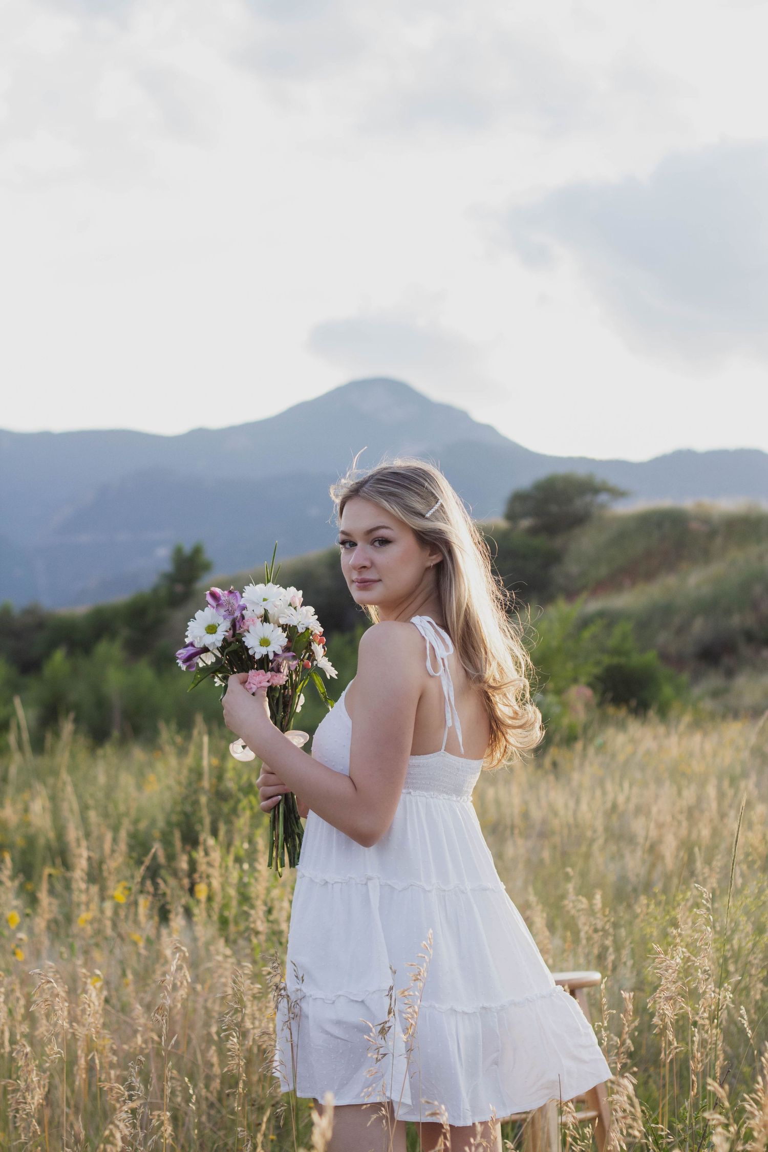 A woman in a white sundress holding a pink and purple bouquet poses in a mountain meadow at sunset. Senior pictures Colorado Springs