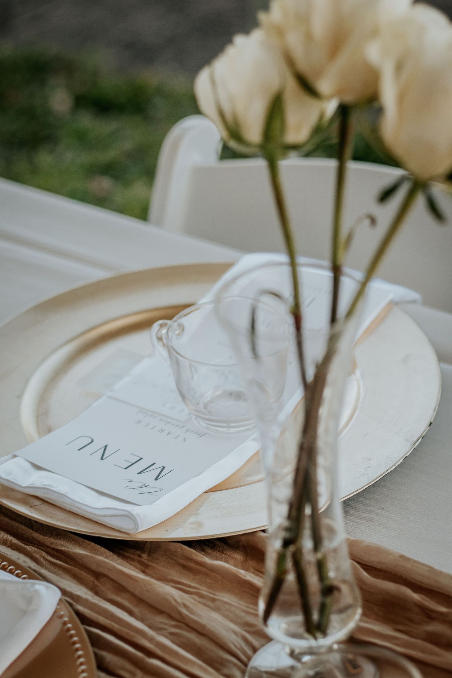 Elegant place setting with white roses in a clear vase and menu card on cream tablecloth.