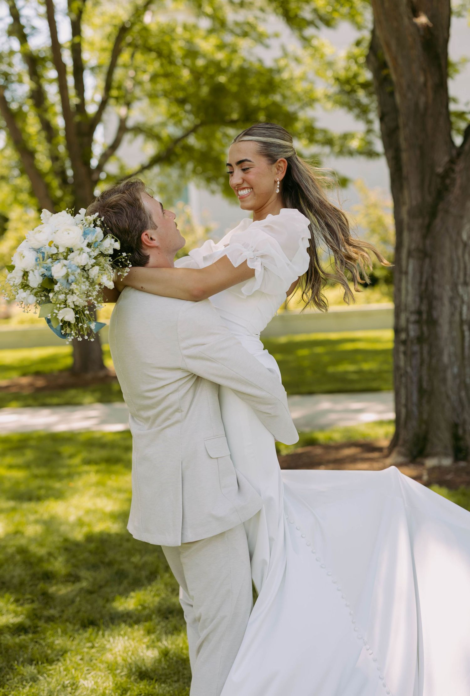 A joyful wedding couple embraces outdoors in a sunlit park with white floral bouquet and formal attire.