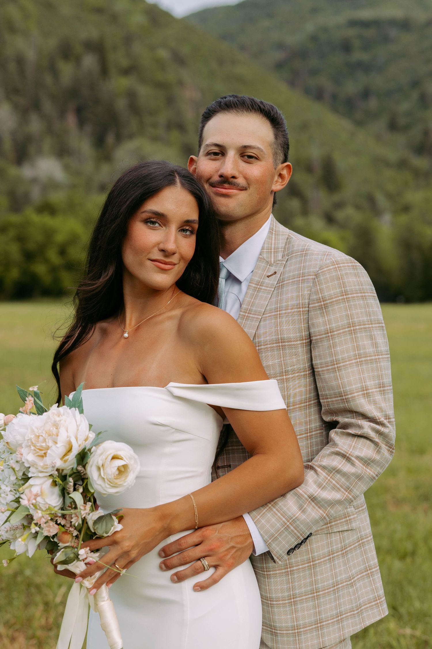 A bride in an off-shoulder white dress holds a blush and white bouquet next to her partner in a plaid suit outdoors.