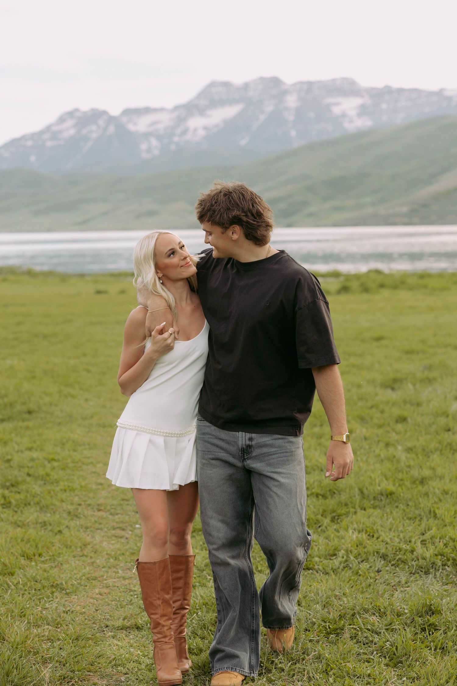 A couple shares a romantic moment on grassy hills with snow-capped mountains and a lake in the background.