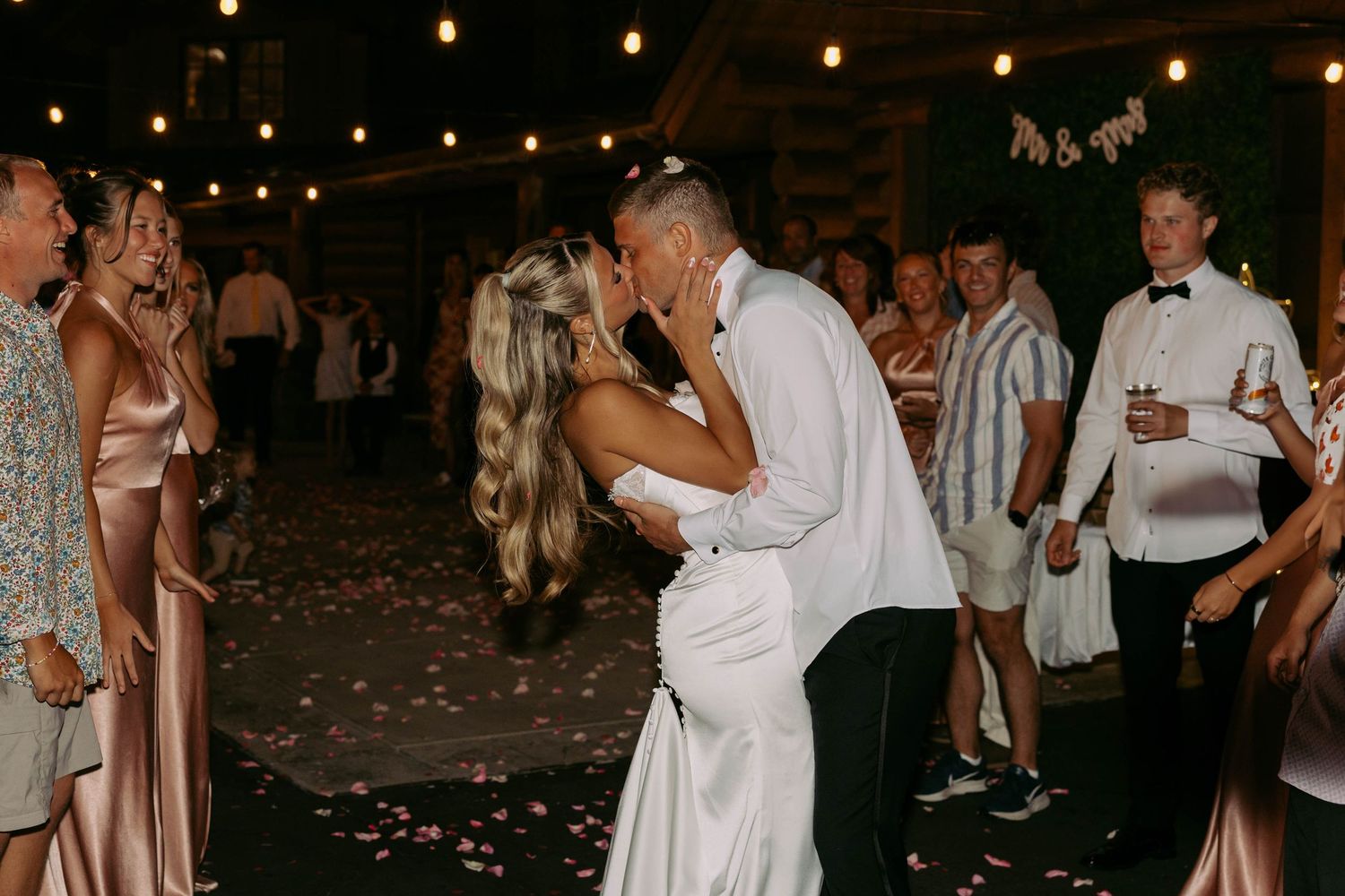 Wedding guests dance and celebrate on a dimly lit dance floor under string lights at an evening reception.