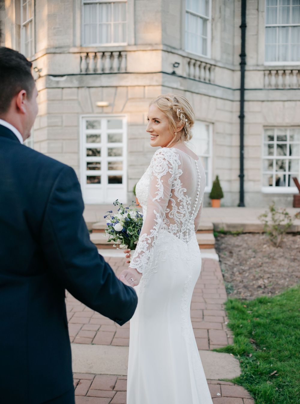 Bride in long-sleeve lace wedding gown looks back while holding bouquet outside elegant stone building.