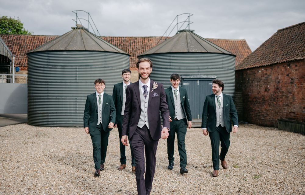 Group of wedding party groomsmen in formal suits walking past farm grain silos on a cloudy day.