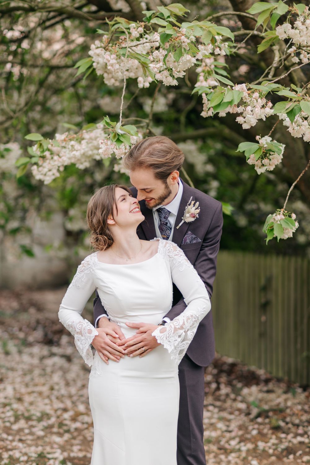 A romantic spring wedding portrait under blooming white cherry blossoms with fallen petals scattered on the ground.