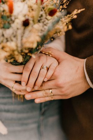 Two hands wearing engagement and wedding rings hold each other with dried flowers in the background.