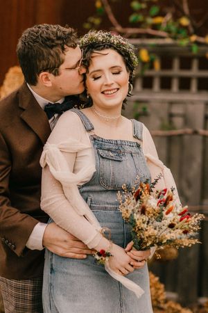 Romantic autumn wedding portrait with couple sharing tender moment while holding dried flower bouquet against wooden fence.