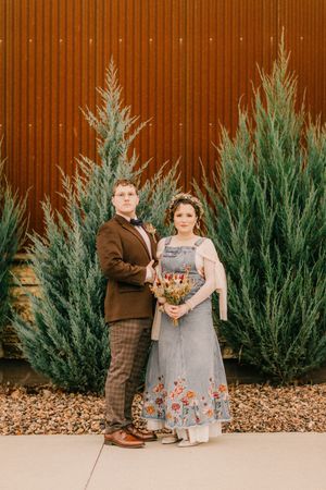 A couple stands in front of tall evergreen trees against a rust-colored wall, wearing vintage-inspired attire and holding a bouquet.