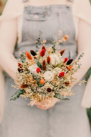A rustic dried flower bouquet with strawflowers, red berries, and delicate white blooms held against a light gray dress.