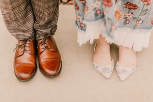 Contrasting wedding shoes with brown leather oxford dress shoes next to delicate white lace flats on beige carpet.